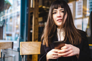 Beautiful young woman with bangs holding smart phone at sidewalk cafe - AMWF00156