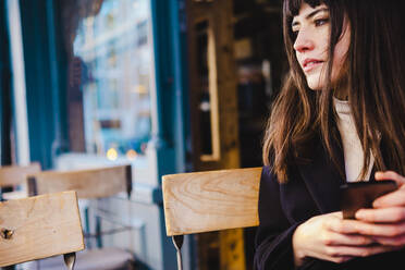 Young woman holding smart phone sitting at sidewalk cafe - AMWF00157