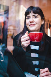 Smiling young woman holding coffee mug looking out through window - AMWF00160