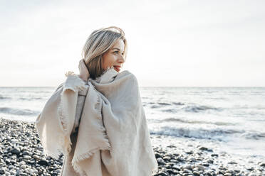 Lächelnde blonde Frau in Decke eingewickelt am Strand - OMIF00463