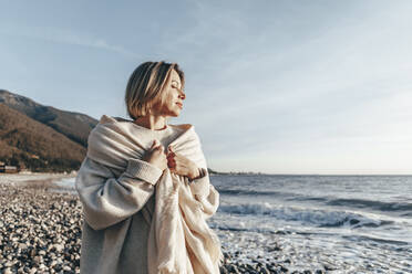 Blonde Frau, eingewickelt in eine Decke, genießt den Sonnenuntergang am Strand - OMIF00466