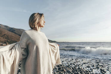 Blonde Frau mit geschlossenen Augen hält eine Decke am Strand - OMIF00467