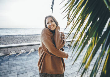 Lächelnde junge Frau mit Pullover an einem Palmenblatt an der Promenade - OMIF00469