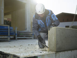 Arbeiter säubert eine Ecke einer Zementplatte mit einem Hammer, der auf der Baustelle kniet - CVF01816