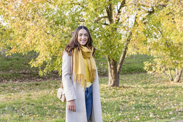 Smiling young woman with yellow scarf standing in autumn park - EIF03139