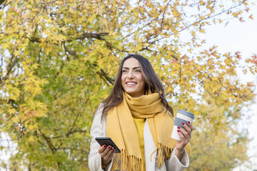 Lächelnde junge Frau mit Smartphone und wiederverwendbarem Kaffeebecher im Herbstpark - EIF03153