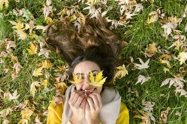 Glückliche junge Frau liegt im Gras und hält Herbstblätter im Park - EIF03158