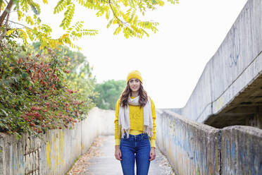 Young woman in yellow jumper and knit hat standing on concrete pedestrian walkway in autumn park - EIF03160