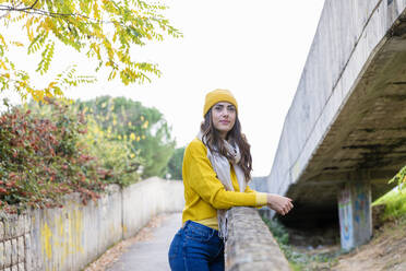 Junge Frau in gelbem Pullover und Strickmütze lehnt an einer Mauer im herbstlichen Park - EIF03163
