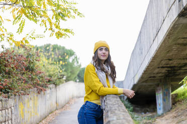 Young woman in yellow jumper and knit hat leaning on wall in autumn park - EIF03163