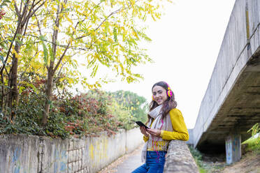 Lächelnde junge Frau hört Musik über Kopfhörer auf einem Fußgängerweg im Herbstpark - EIF03164