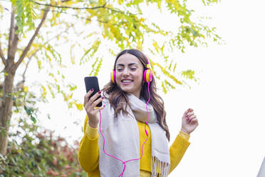 Smiling young woman enjoying music through headphones in autumn park - EIF03166
