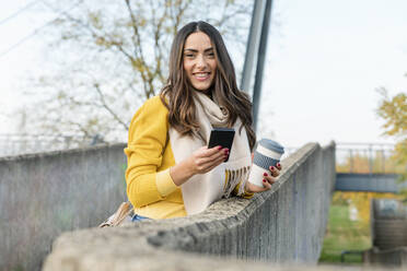 Smiling young woman with smart phone and reusable coffee cup leaning on wall - EIF03169