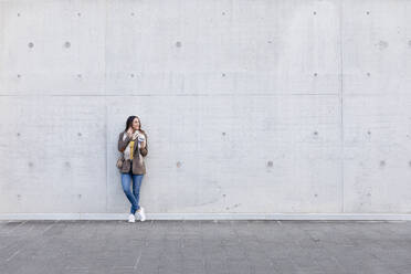 Junge Frau mit wiederverwendbarem Kaffeebecher, die vor einer grauen Wand mit ihrem Smartphone telefoniert - EIF03171