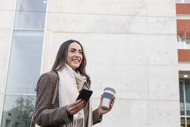 Smiling young woman with smart phone and reusable coffee cup in front of building - EIF03173