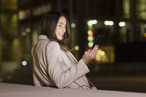 Junge Frau, die ein Smartphone benutzt und sich nachts an die Wand lehnt - EIF03176