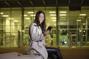 Glückliche Geschäftsfrau, die nachts vor einem Bürogebäude ein Mobiltelefon benutzt - EIF03179