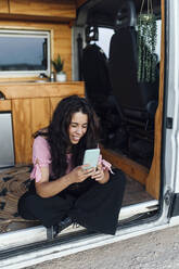 Glückliche junge Frau, die ihr Smartphone in einem auf einem Feldweg geparkten Lieferwagen benutzt - EGHF00332
