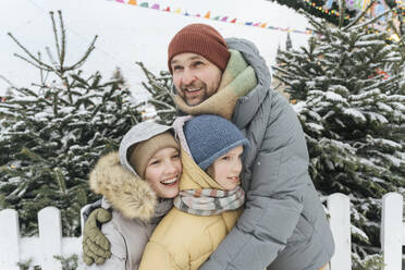 Smiling father embracing children in warm clothing at backyard - EYAF01871