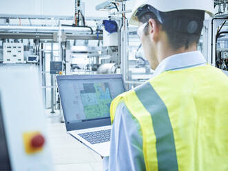 Junger Ingenieur in Arbeitsschutzkleidung mit Laptop in einer Fabrik - CVF01844
