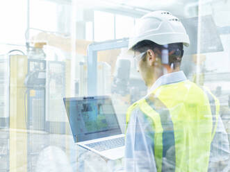 Junger Ingenieur in Arbeitsschutzkleidung mit Laptop durch eine Glaswand in einer Fabrik gesehen - CVF01853