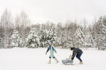 Vater, der seine Tochter auf einem Schlitten durch eine Frau schiebt, die im Winter im Schnee spazieren geht - EYAF01878