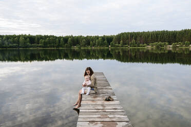 Mother spending leisure time with baby girl by lake on jetty - EYAF01895