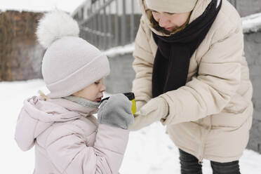 Mother feeding daughter through cup in winter - SEAF00406