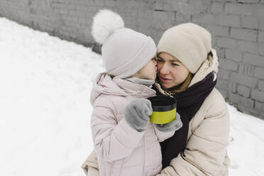 Girl holding cup while kissing mother on cheek in winter - SEAF00407