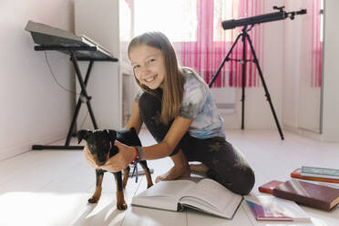 Lächelndes Mädchen mit Büchern, das einen Miniaturpinscher-Hund zu Hause hält - EYAF01897