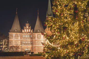 Germany, Schleswig-Holstein, Lubeck, Illuminated Holstentor gate at night with glowing Christmas tree in foreground - KEBF02205