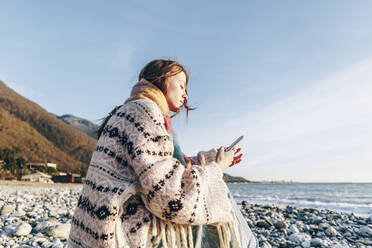 Teenager-Mädchen, das am Strand sitzend ein Smartphone benutzt, Gagra, Abchasien - OMIF00474