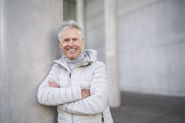 Glücklicher älterer Mann mit verschränkten Armen an der Wand lehnend - PNEF02618