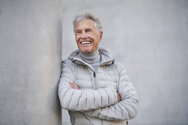 Lächelnder älterer Mann mit verschränkten Armen an einer grauen Wand - PNEF02619