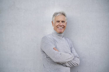 Happy senior man with arms crossed near gray wall - PNEF02624