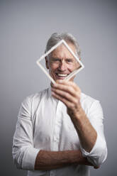 Smiling businessman looking through frame in front of gray background - PNEF02665