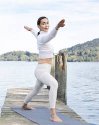 Smiling woman with arms outstretched exercising on jetty by lake - JBYF00101