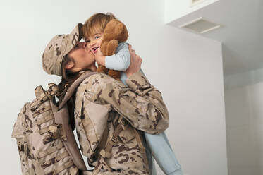 Seitenansicht der glücklichen weiblichen Soldat in Uniform mit Rucksack hält küssen niedlichen Sohn im Schlafzimmer - ADSF33431