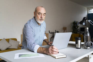 Geschäftsmann schreibt in einen Notizblock und sitzt mit einem Laptop am Schreibtisch - GIOF14740