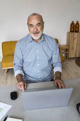Lächelnder älterer Freiberufler mit Laptop am Schreibtisch im Heimbüro sitzend - GIOF14745