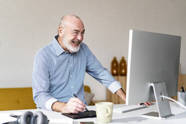 Cheerful freelancer using graphic tablet and computer at desk in home office - GIOF14764