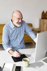 Cheerful senior businessman with graphic tablet looking at computer in home office - GIOF14765