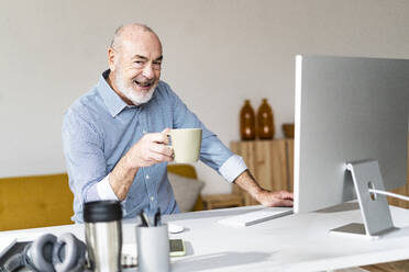 Smiling freelancer holding coffee mug sitting at desk - GIOF14771