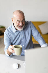 Glücklicher älterer Freiberufler, der eine Kaffeetasse hält und im Heimbüro arbeitet - GIOF14773