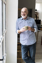 Happy businessman holding coffee mug leaning on wall at home - GIOF14796