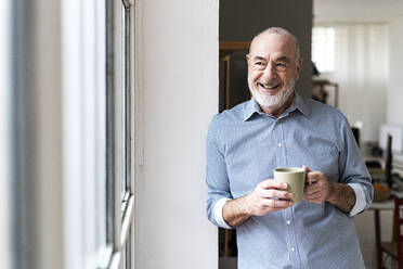 Happy senior freelancer holding coffee cup looking through window - GIOF14797