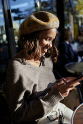 Lächelnde Frau mit Baskenmütze und Smartphone an einem sonnigen Tag - KIJF04372