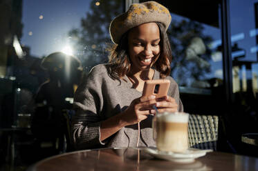 Lächelnde Frau mit Baskenmütze, die eine Kaffeetasse auf dem Tisch eines Cafés fotografiert - KIJF04374