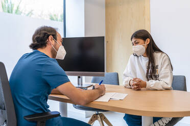 Arzt im Gespräch mit einem am Schreibtisch sitzenden Patienten im Krankenhaus während einer Pandemie - DLTSF02644