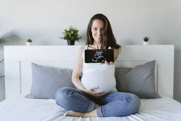 Smiling pregnant woman showing ultrasound scan through smart phone at home - EBBF05219